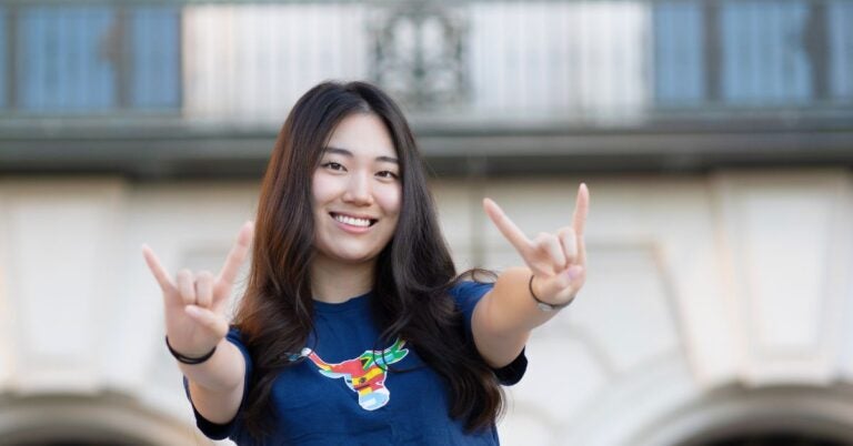 A young Asian woman smiles while wearing a navy blue Texas Global tshirt and making UT hook'em hand signs outdoors.