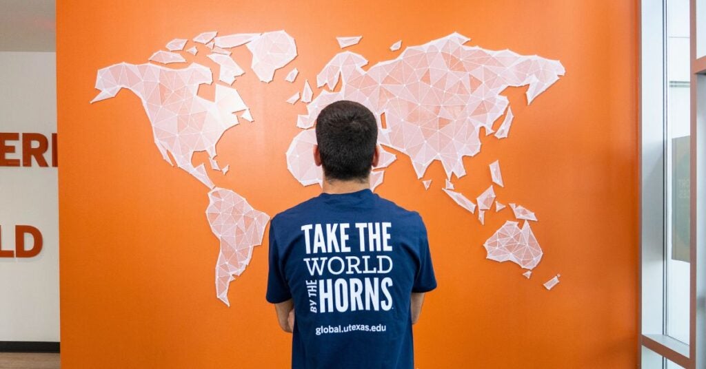 A person stands facing a geometric world map on an orange wall, wearing a blue shirt with the phrase "Take the World by the Horns." The tone is inspirational.