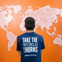 A person stands facing a geometric world map on an orange wall, wearing a blue shirt with the phrase "Take the World by the Horns." The tone is inspirational.