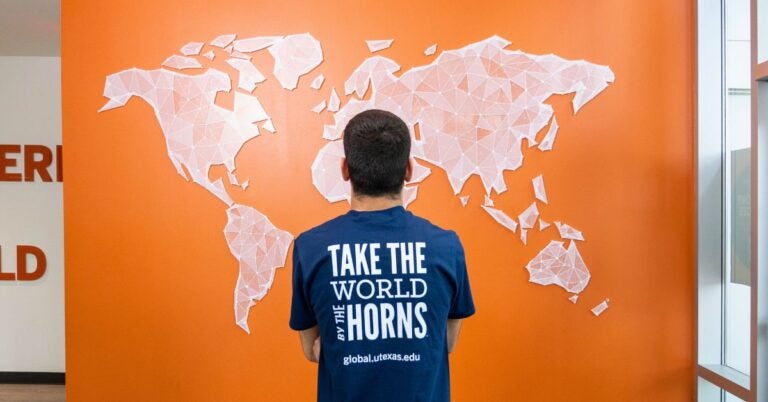A person stands facing a geometric world map on an orange wall, wearing a blue shirt with the phrase "Take the World by the Horns." The tone is inspirational.