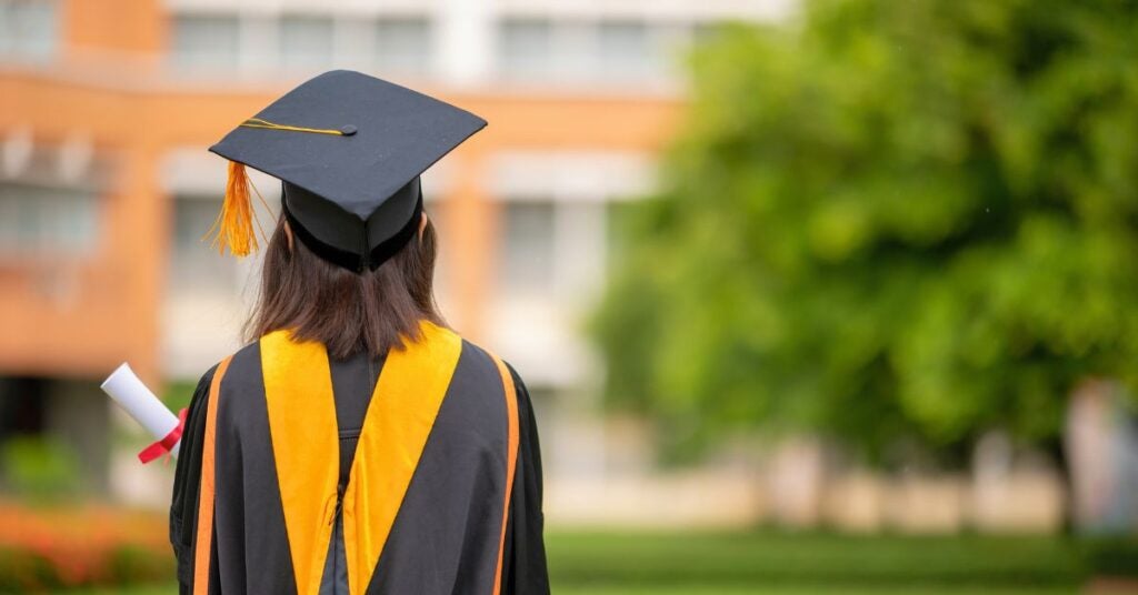 The image shows the back view of a graduate wearing a black cap and gown with gold accents. The graduate stands outdoors, holding a white rolled diploma tied with a red ribbon in the left hand.