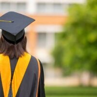 The image shows the back view of a graduate wearing a black cap and gown with gold accents. The graduate stands outdoors, holding a white rolled diploma tied with a red ribbon in the left hand.