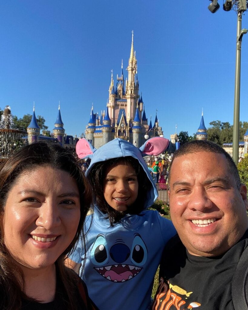 Family of three smiling in front of a Disney castle, child in a Stitch hoodie. Sunny day, clear blue sky, capturing joy and excitement.