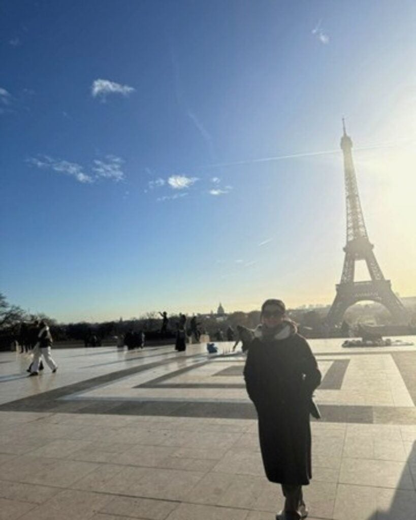 The image shows a person standing on a large plaza with the Eiffel Tower visible in the background. The scene is set on a clear day with a bright, blue sky and some scattered clouds. The sunlight is casting long shadows across the plaza.