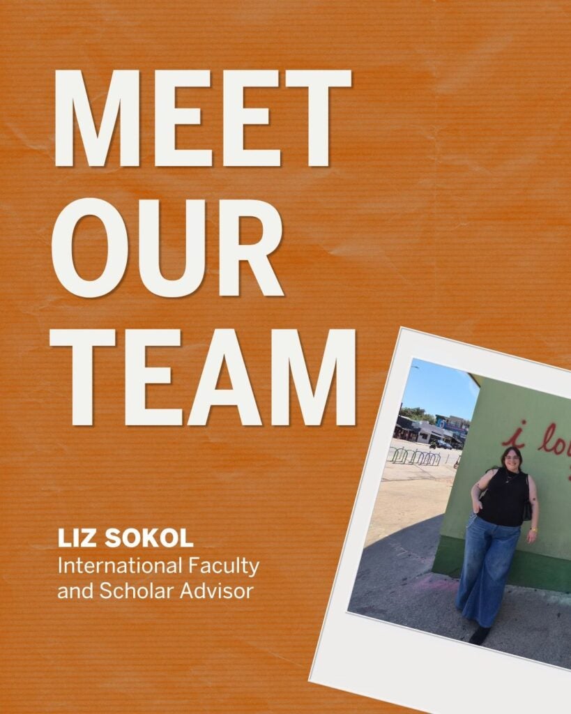 The image features a promotional graphic with a textured orange background. On the left, large bold white text reads "MEET OUR TEAM." Below this, in smaller white text, it says "LIZ SOKOL International Faculty and Scholar Advisor." On the right side of the image, there is a tilted photograph of a person standing in front of a green wall with cursive red lettering. The person is wearing a black top and wide-legged jeans. The photo is designed to resemble a Polaroid picture.