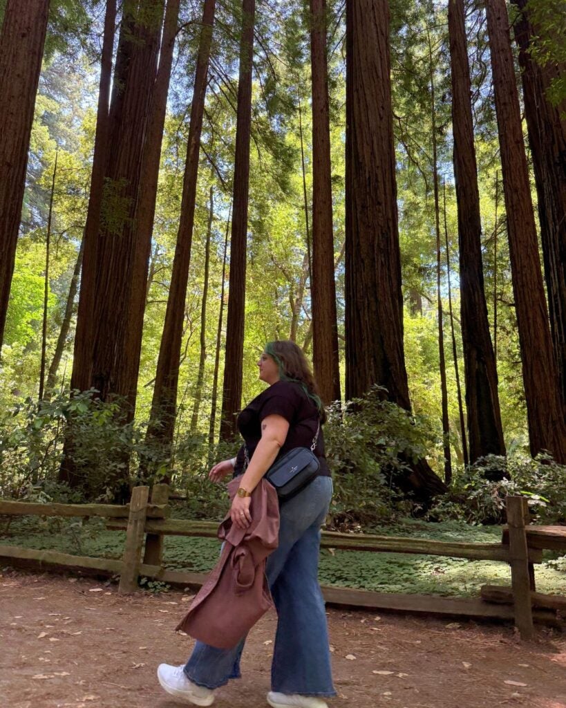 Woman walking along a dirt path in a dense forest of tall redwood trees on a sunny day.