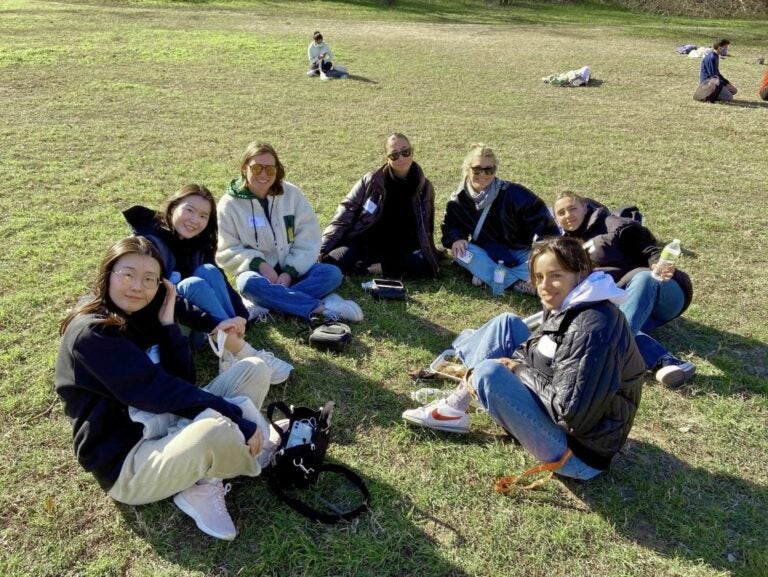 Group of seven female friends sitting in a circle on a grassy field enjoying the company and a sunny day outdoors.