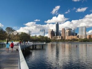 People walking along a lakeside boardwalk with Austin's city skyline under a partly cloudy blue sky in the background.