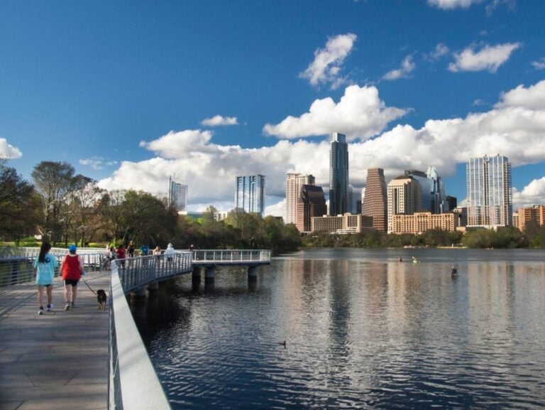 People walking along a lakeside boardwalk with Austin's city skyline under a partly cloudy blue sky in the background.
