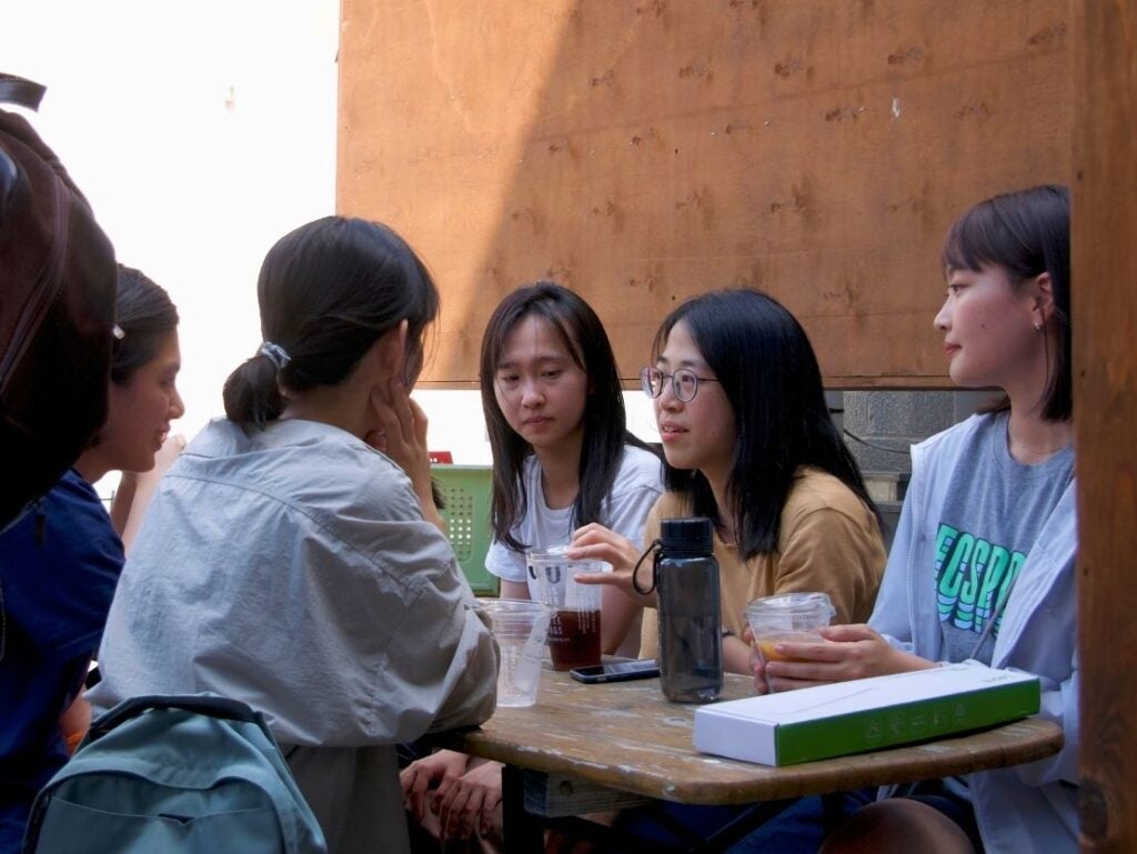 Five young women engaged in a focused conversation around a wooden table with drinks and a book.