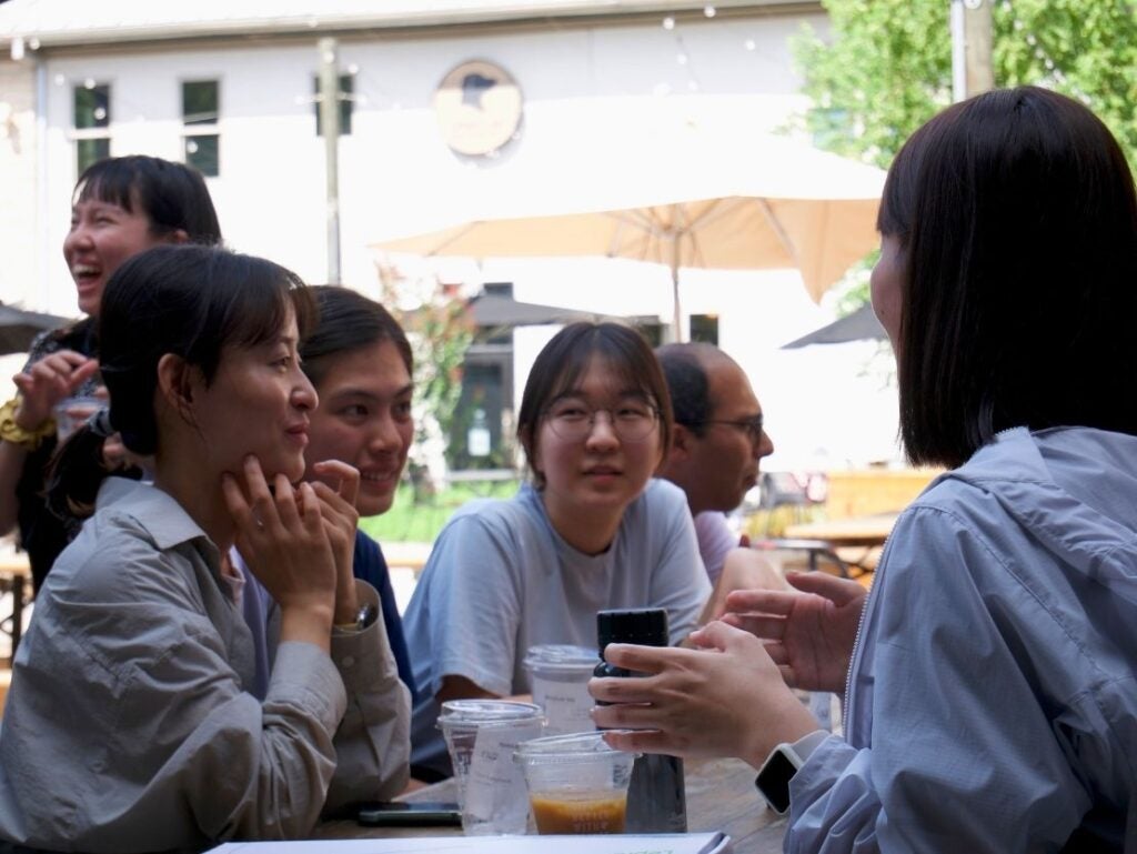 Group of young adults engaged in lively conversation around a table on a sunny day.