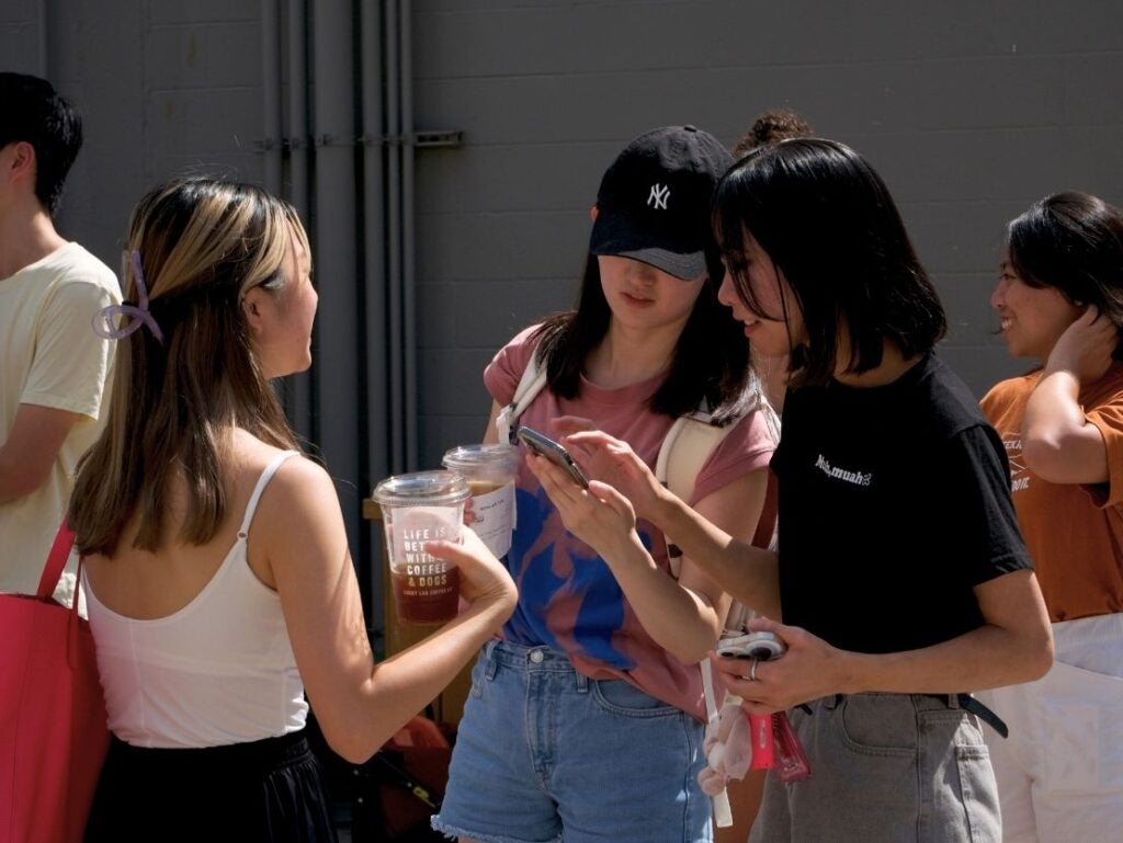 Three young women stand outdoors chatting, one holding iced coffee cups while another shows something on her phone.