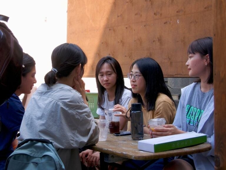 Five young women engaged in a focused conversation around a wooden table with drinks and a book.