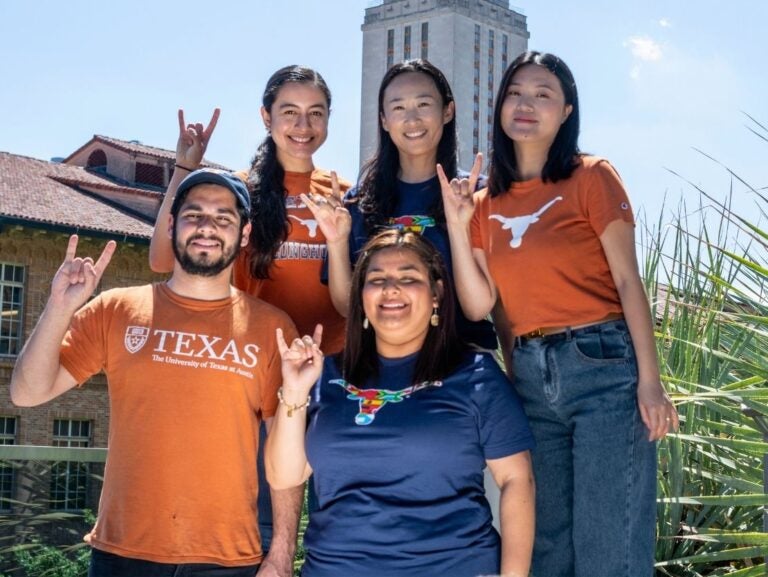 Five University of Texas students outdoors making the Hook 'em Horns hand sign with a campus building in the background.