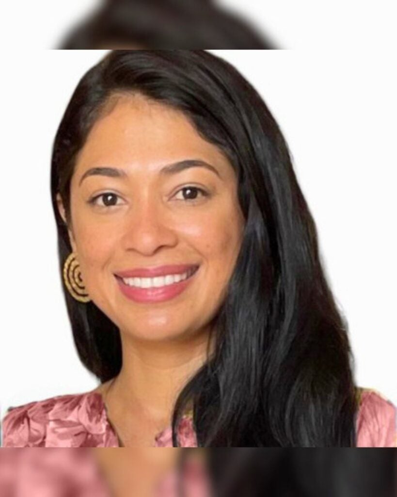 Smiling woman with long black hair wearing gold earrings and a pink blouse against a white background.