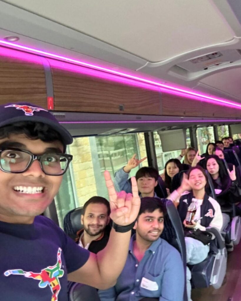 Group of diverse young adults smiling and making hand signs while seated inside a bus with purple ambient lighting.