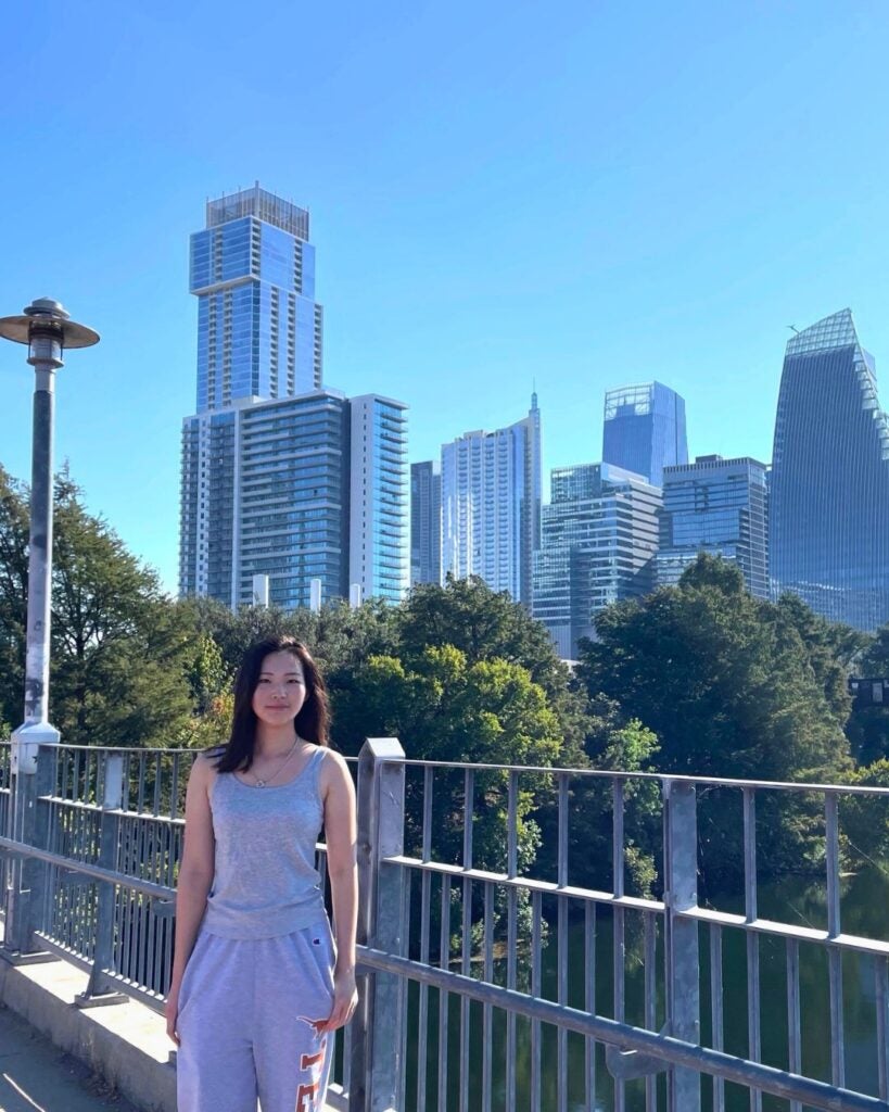 Young woman in casual gray outfit standing on a bridge with a city skyline and green trees in the background under a clear blue sky.
