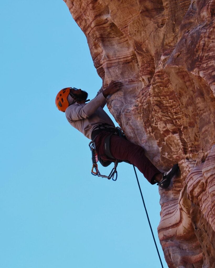 Man wearing an orange helmet and climbing gear scaling a steep, textured red rock face against a clear blue sky.