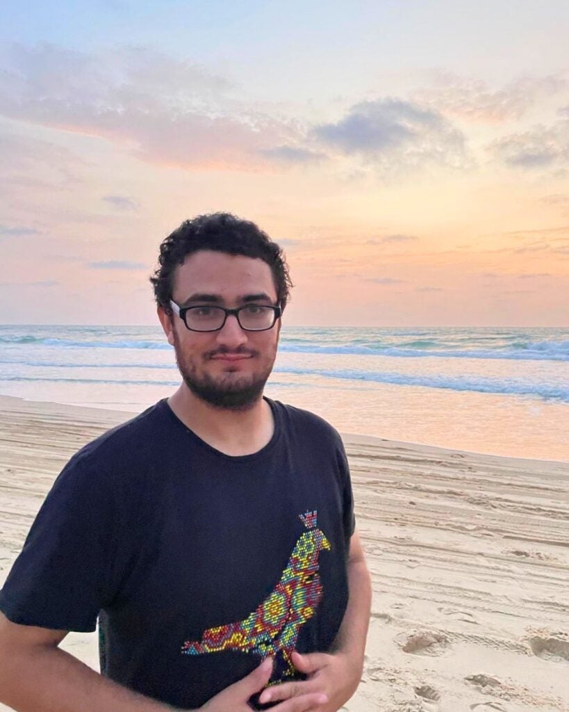 Young man with glasses standing on a sandy beach at sunset with gentle waves in the background.