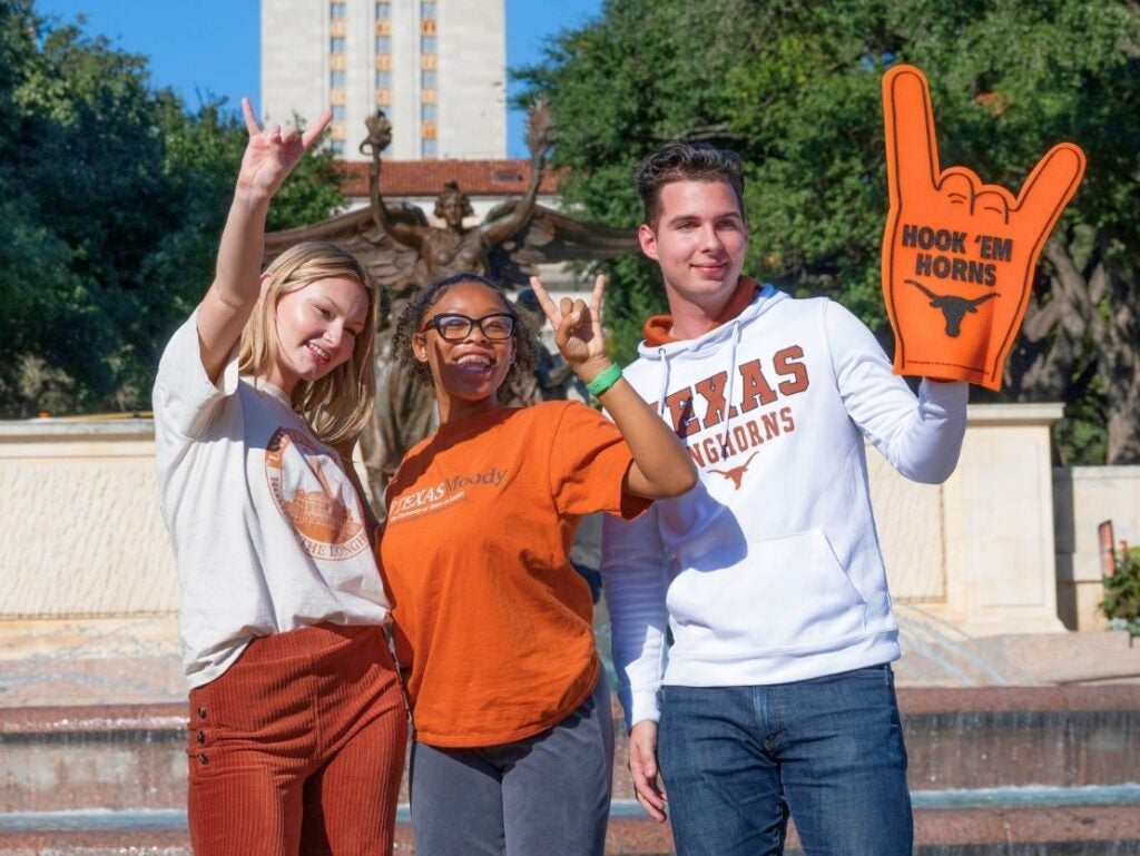 Three University of Texas students posing in front of the UT Tower, making the Hook 'Em Horns hand sign, one holding a large foam hand with the slogan.