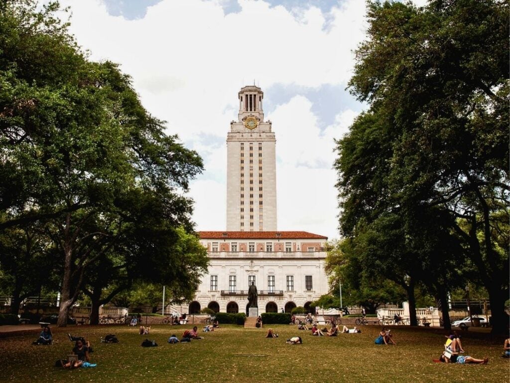 The University of Texas Tower rises above a green lawn where students relax under large trees on a sunny day.