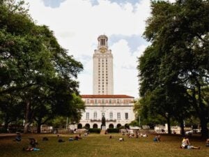 The University of Texas Tower rises above a green lawn where students relax under large trees on a sunny day.