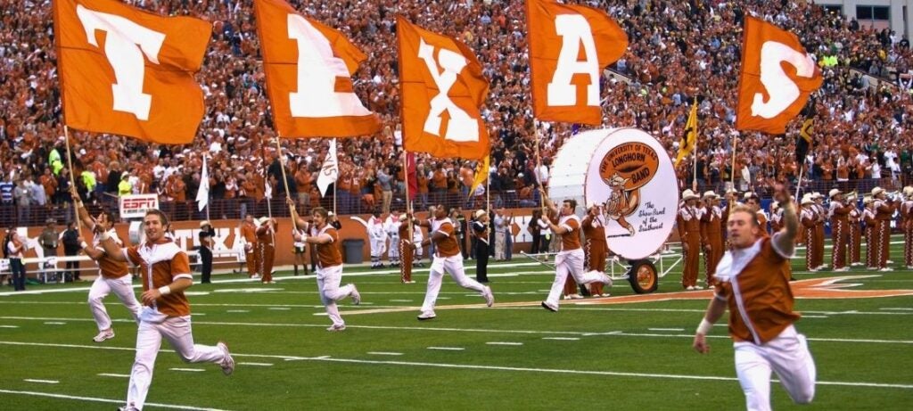 Texas Longhorns marching band performs on a football field as giant orange TEXAS flags wave before a packed, cheering stadium crowd.