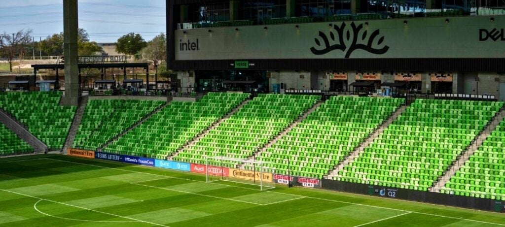 Empty stadium seating with bright green chairs surrounding a well-maintained soccer field under a clear sky.