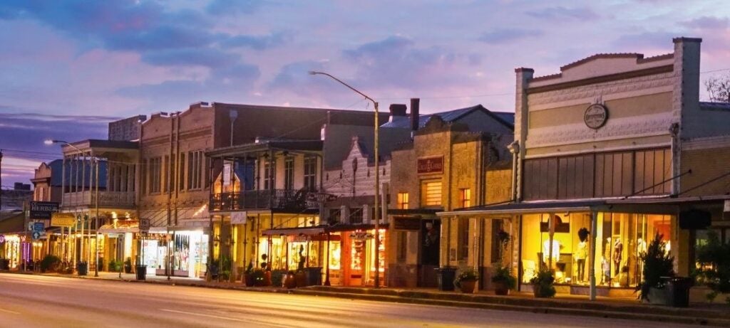 Historic downtown street at dusk with warmly lit storefronts and a colorful sky overhead.