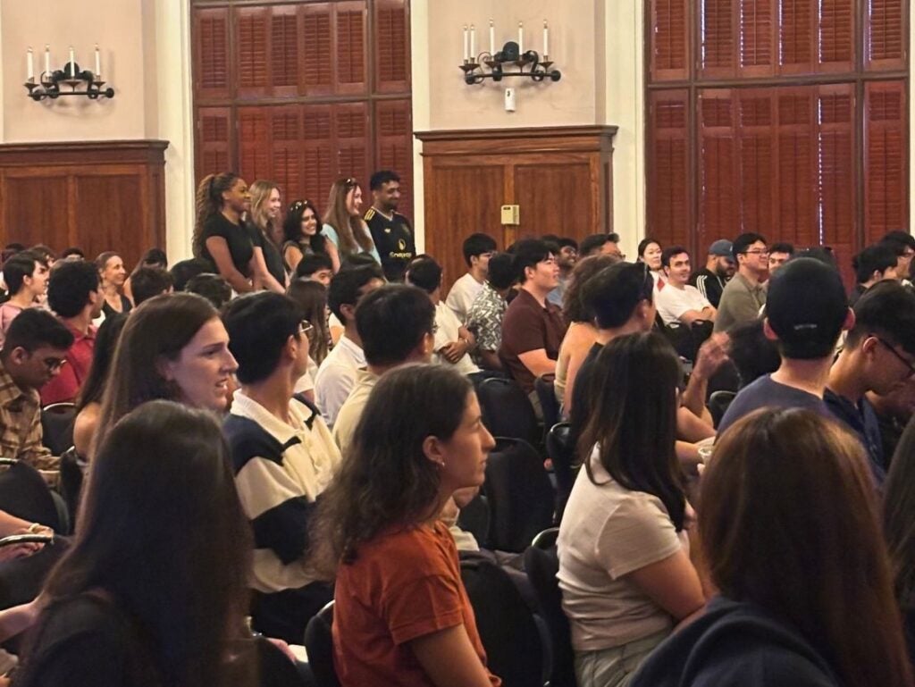 A diverse group of people attentively listening in a wood-paneled room with large windows and wall sconces.