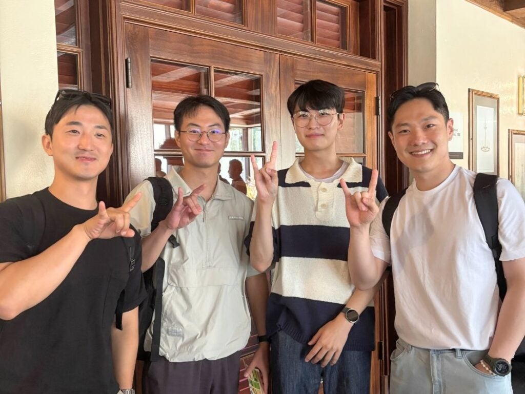 Four young men standing indoors smiling and making the Texas Longhorns hand sign in front of wooden doors.