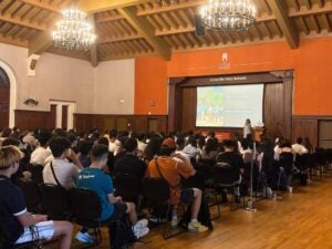 Audience attentively listening to a speaker presenting slides in a large, wood-paneled ballroom with chandeliers.