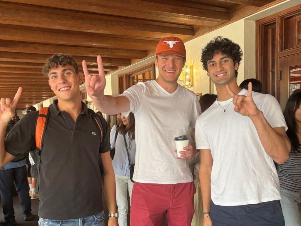 Three young men smiling and making the Texas Longhorns hand sign in a wooden-beamed hallway, one wearing a burnt orange cap and holding a coffee cup.
