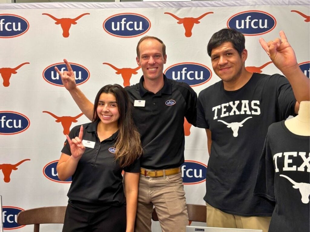 Three people smiling and making the Texas Longhorns hand sign in front of a UFCU and Texas Longhorns logo backdrop.