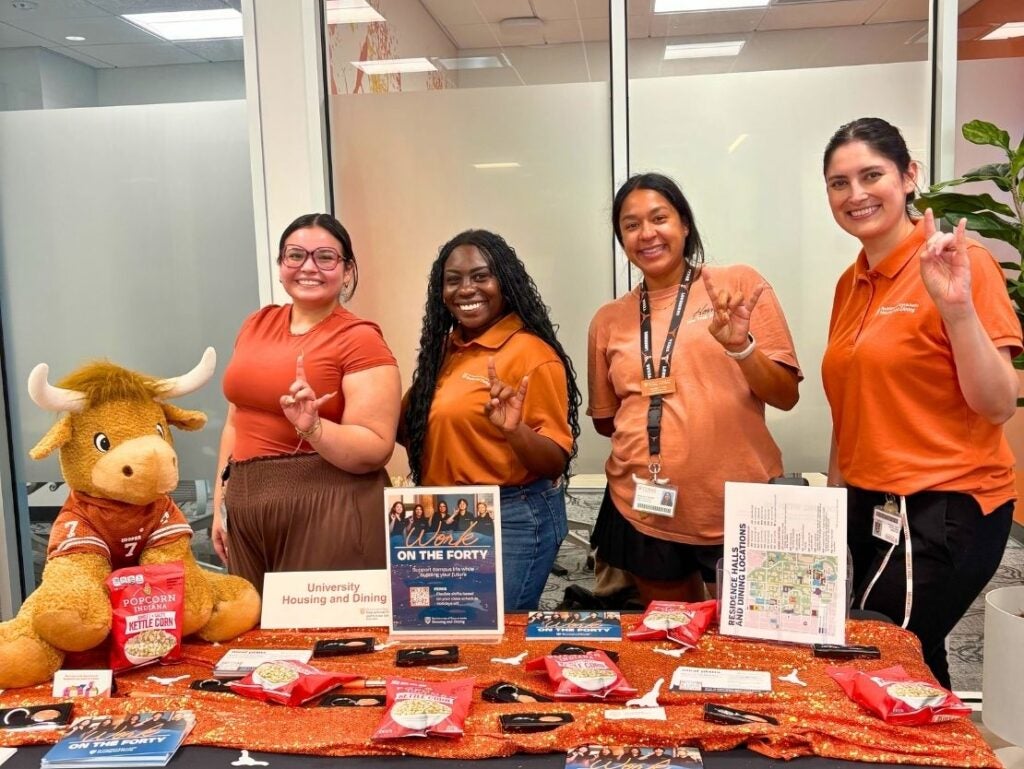 Four women in orange shirts smiling and posing behind a university housing and dining information table with a stuffed longhorn mascot.