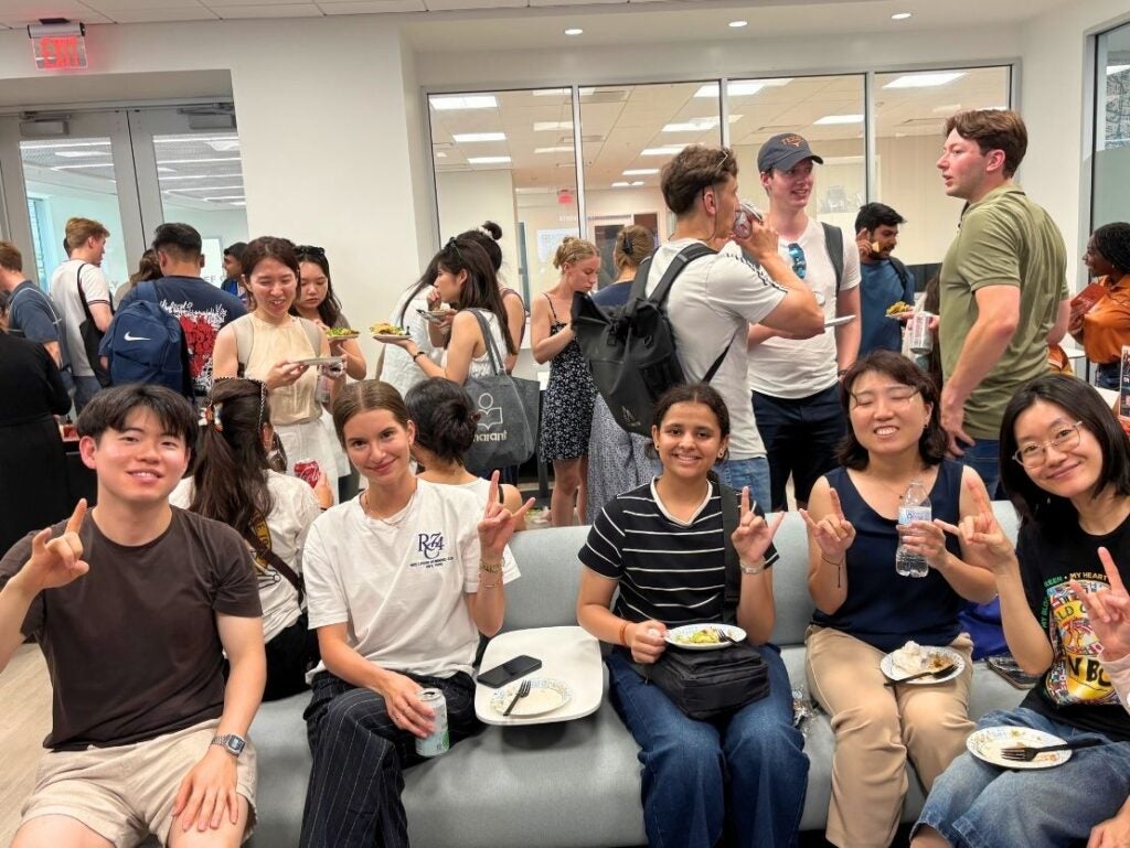 Group of diverse young adults sitting and standing in a bright room, enjoying food and socializing at a casual gathering.