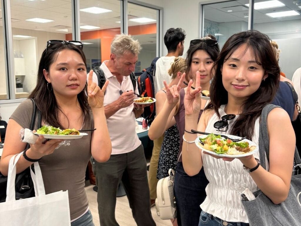 Three young women holding plates of salad and making hand gestures at an indoor social gathering with other attendees in the background.