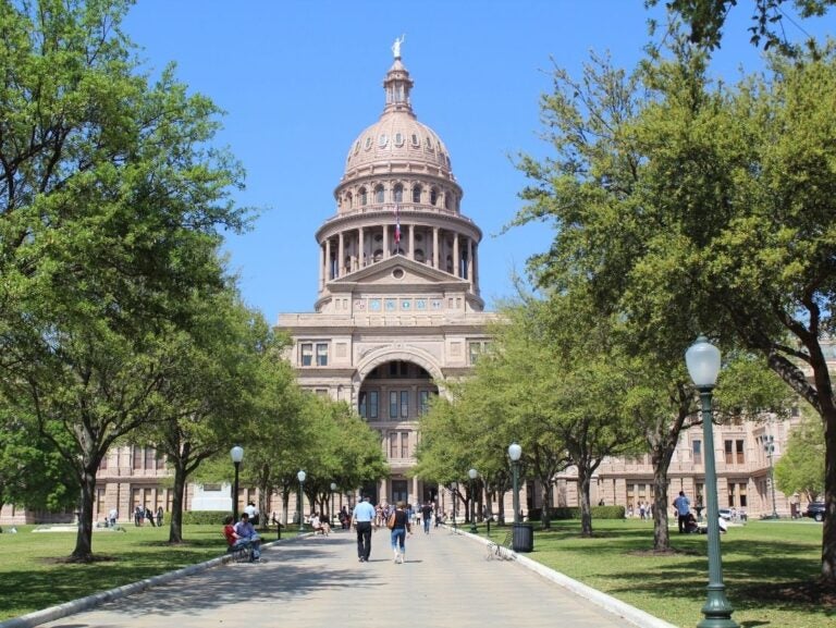 View of the Texas State Capitol building with a clear blue sky and people walking along the tree-lined pathway leading to its entrance.