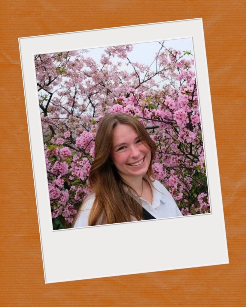 Young woman with long brown hair smiling in front of vibrant pink cherry blossoms on a sunny day.