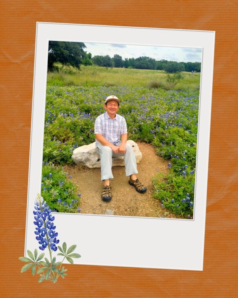 Man in a plaid shirt and hat sitting on a rock surrounded by blooming bluebonnets in a green field under a cloudy sky.