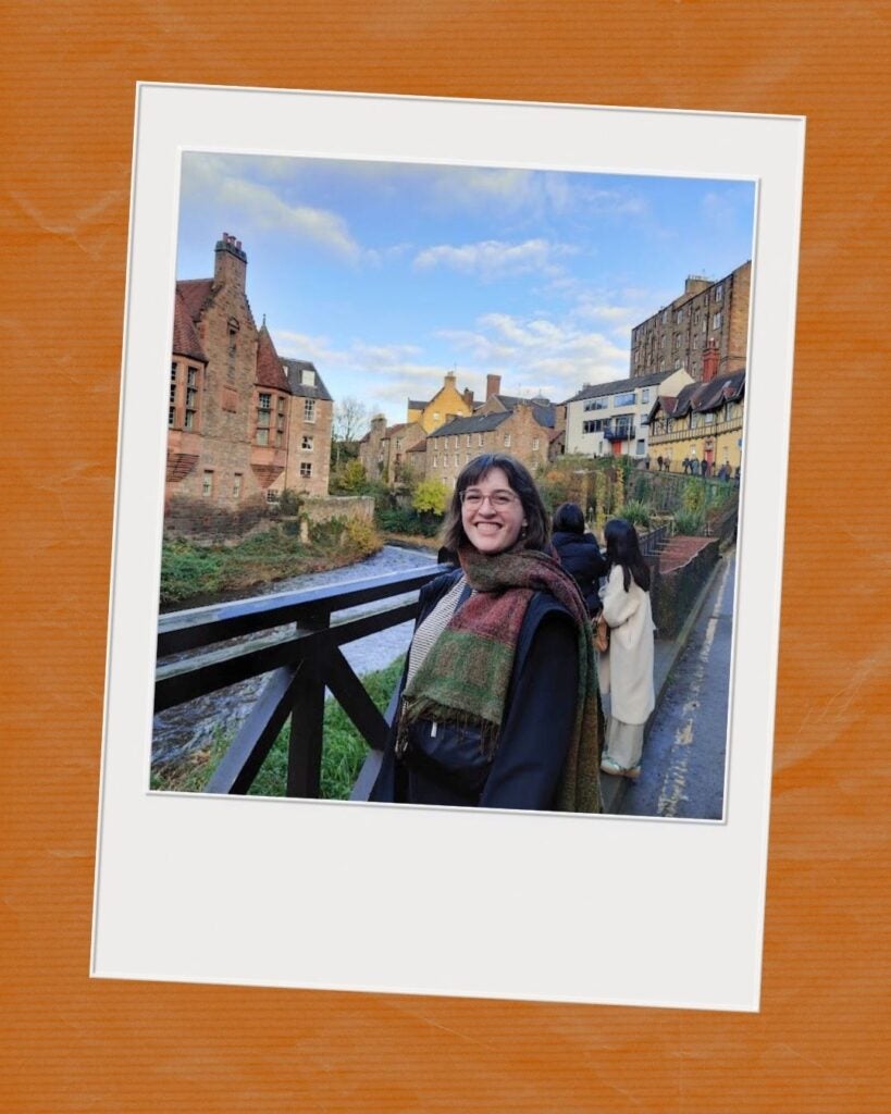 Smiling woman wearing a colorful scarf stands on a wooden bridge overlooking a river with historic stone buildings under a blue sky.
