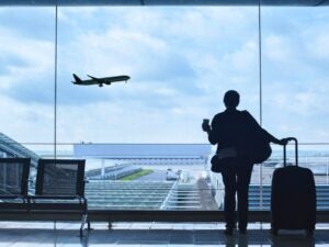 Traveler with suitcase and coffee cup watches airplane take off through large airport window.