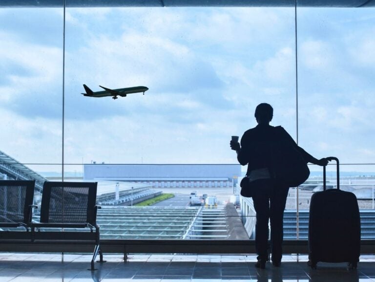 Traveler with suitcase and coffee cup watches airplane take off through large airport window.